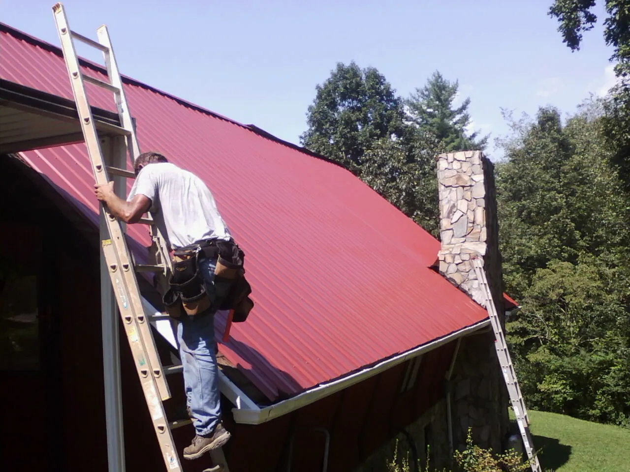 Roofer on a ladder beside a red metal roof and stone chimney — Soffit & Fascia Repair by Roofing Brothers in Placitas